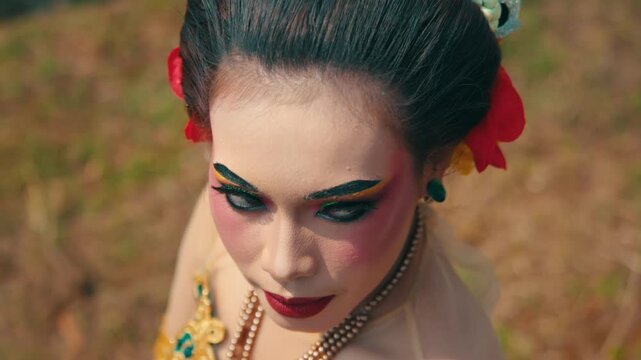 High angle view of an Indonesian dancer's face with heavy traditional makeup and red flowers, looking down and then glancing to the side.
