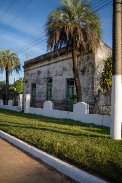 View of a weathered historic building with green doors and windows, framed by palm trees and a white fence in Guaviravi, Corrientes Province, Argentina.