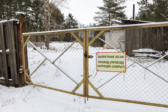 A rusty chain link gate with a yellow Russian restricted area sign blocks access to a snowy rural yard. Russian text means: Restricted area! pedestrian and vehicular access prohibited and closed