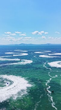 Various watercraft are moving across the ocean, creating waves and patterns in the water. This scene takes place under a clear blue sky with clouds.