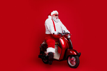 Obraz premium Santa Claus in red suit riding a shiny red scooter for Christmas celebration and holiday fun, playful festive portrait with white beard and sunglasses on vibrant red background