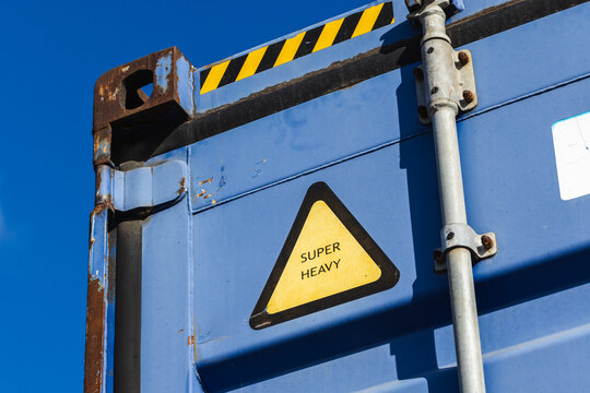 Close-up of a blue shipping container door featuring a yellow Super Heavy warning triangle, rusted metal hardware and safety stripes against a clear sky