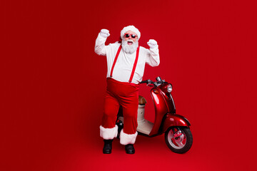 Obraz premium Santa Claus in red sunglasses cheering beside a vintage red scooter on a bold red background for playful Christmas holiday celebration