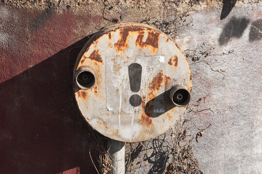 A rusted circular electrical distribution box with an exclamation mark mounted on a sunlit, weathered wall, surrounded by dried vines and peeling paint, evoking industrial decay and urban texture