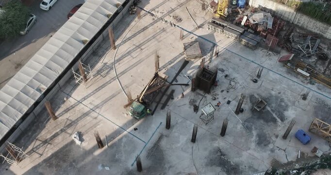 Aerial perspective shows a crane truck unloading steel reinforcements on a construction site, while workers assemble the structural columns.
