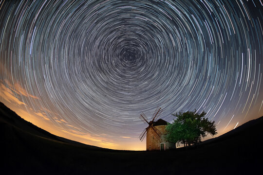 View of a historic windmill under a night sky filled with circular star trails in a rural landscape in Brno-City District, South Moravian Region, Czechia.