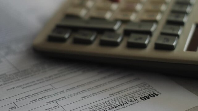 A taxpayer performs calculations on a calculator next to the U.S. Form 1040 tax return document. The close-up shot illustrates financial reporting, accounting, and annual tax filing workflows.