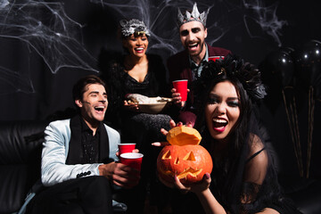 Obraz premium Celebrating Halloween with asian woman holding carved pumpkin near multicultural friends in costumes