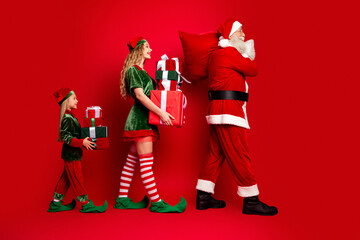 Obraz premium Santa Claus and two festive elves carrying wrapped Christmas presents in a cheerful holiday procession against a red background