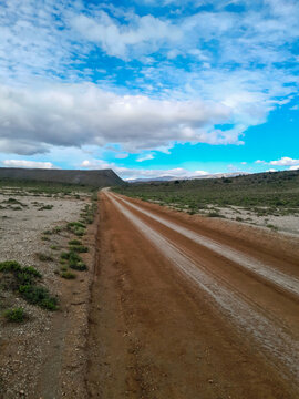 View of a dirt road winding through the semi-arid landscape of Anysberg Nature Reserve with distant hills under a cloudy blue sky in Anysberg, Western Cape, South Africa.