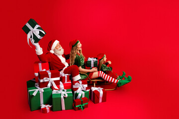 Obraz premium Santa Claus and two cheerful elves with wrapped Christmas gifts on a sled against a bright red background celebrating holiday joy