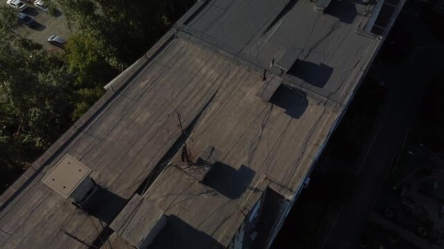 Aerial view of a rooftop with vents and pipes. Sunlight casts shadows across weathered surfaces. Person stands near edge, looking down at street below. Urban setting with parked cars and green trees