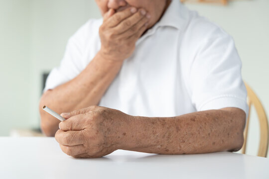 70s senior southeast asian man holding a cigarette at home, feeling sick and regretful about smoking indoors, concept image for quit smoking awareness
