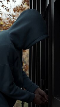 Criminal Prowler Tries Opening Glass Door On Private Residential Terrace, Seeking Access Inside. Mysterious Intruder In Dark Sweatshirt Works Handle Secretly Against Overcast Fall Backdrop.