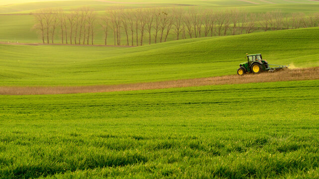 View of a green tractor working in a lush green field with rolling hills in the background in Moravia, Brno-City District, South Moravian Region, Czechia.