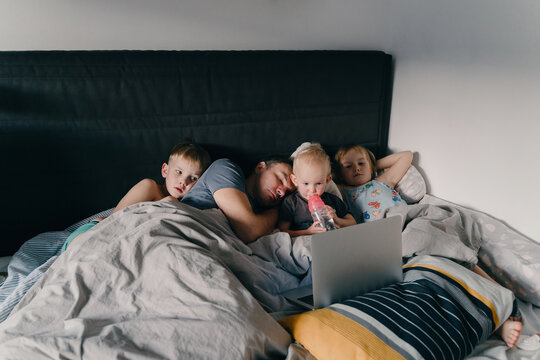 Three little children are watching a laptop while their exhausted father is sleeping beside them in a cozy bed. This authentic family lifestyle concept highlights modern parenting struggles.