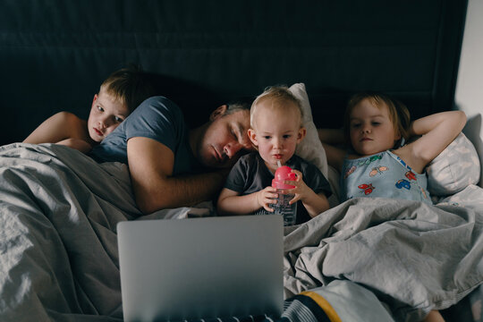 Three little children are watching a laptop while their exhausted father is sleeping beside them in a cozy bed. This authentic family lifestyle concept highlights modern parenting struggles.
