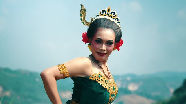 Medium shot of a beautiful female dancer wearing a green traditional outfit, smiling warmly at the camera in a rocky nature park.