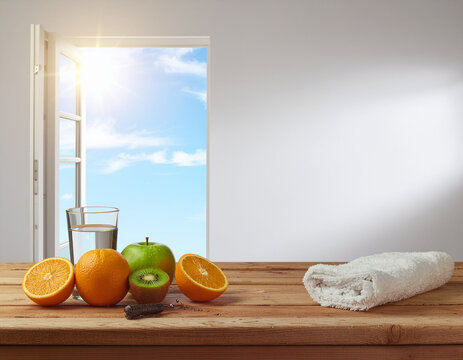 healthy morning wellness concept featuring fresh oranges, green apple, kiwi, a glass of water, and a white towel on a wooden table beside an open window with bright sunshine and blue sky