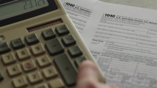A person presses buttons on a calculator while calculating individual income tax next to a 1040 form. An accountant performs a financial audit and plans the personal budget on an office desk