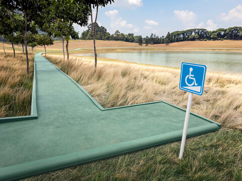 green paved path winds through a landscape next to a calm lake, blue wheelchair accessibility sign points toward the trail, tall, dry grass, scattered trees, and rolling hills under partly cloudy sky