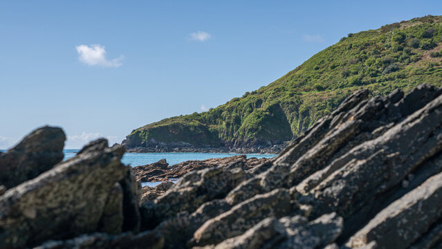 View of the rugged coastline along the South West Coastal Path overlooking Portnadler Bay with dark rocks and green hills under a clear blue sky in Looe, Cornwall, United Kingdom.