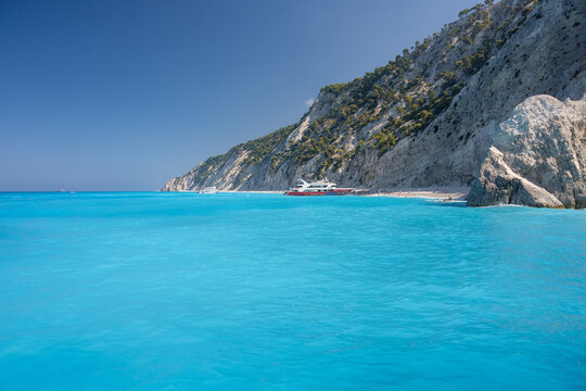 View of the turquoise waters of the Ionian Sea at a beach with steep white cliffs and a passenger boat docked near the shore in Lefkada, Greece.