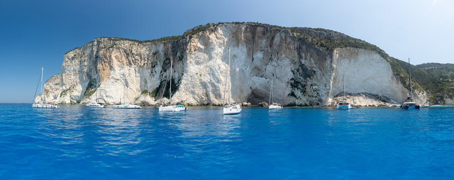 View of sailboats anchored in the turquoise Ionian Sea in front of towering white limestone cliffs on the coast of Lefkada, Greece.