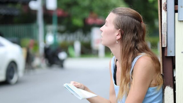 Exhausted and lost female tourist resting on a Chiang Mai street and fanning herself with a map during a hot summer day in Thailand while traveling solo