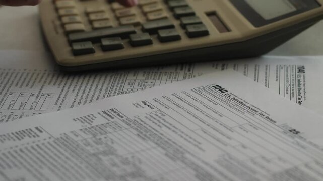 A person uses a calculator to calculate taxes and fill out the 1040 individual income tax return form. Paper financial documents lie on the table during budget calculation and financial audit