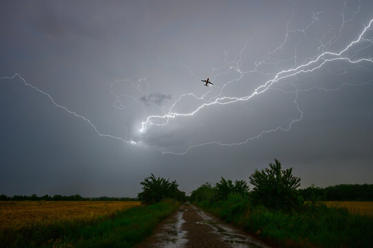 View of a small airplane flying through a dark, stormy sky illuminated by a massive lightning bolt above a rural dirt road in Bucharest, Bucharest, Romania.
