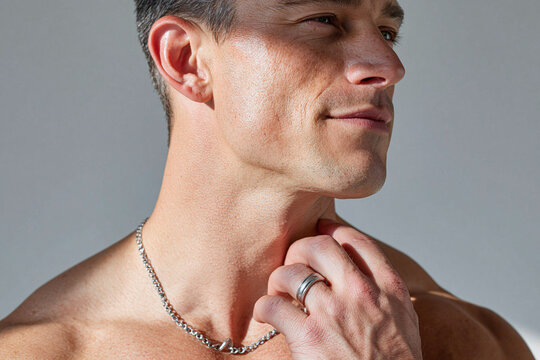 Bare chested man with sunlight grazing collarbone and male fingers lightly touching neck, wearing chain necklace and wedding ring, contemplative expression, warm natural skin texture