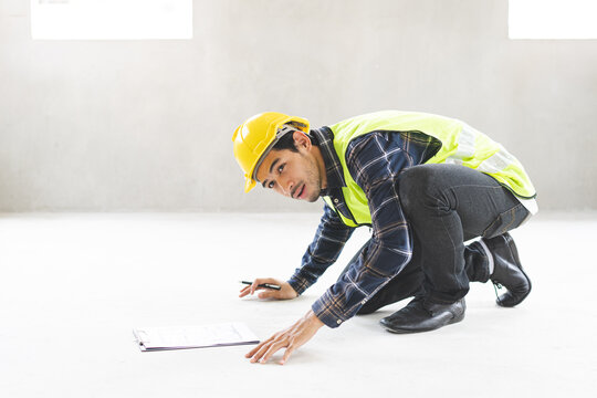 Asian male construction engineer or inspector in safety uniform checking concrete floor quality and writing on clipboard checklist at home building renovation site project.
