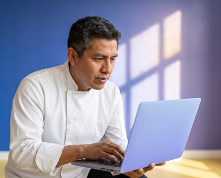 Absorbed chef in white jacket holding open laptop and typing against vivid blue wall