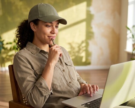 Reflective field worker in olive cap holding pen to lips using laptop near plant