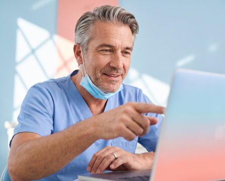 Confident doctor in blue scrubs with lowered mask pointing at laptop screen against pastel wall