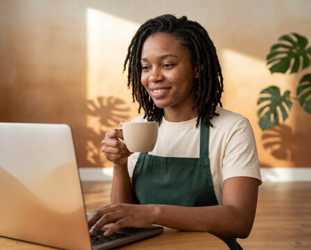 Serene cafe owner in green apron sipping from mug while working on laptop near monstera
