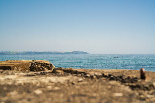 View of the calm sea and horizon with a small boat, framed by a weathered stone structure in the foreground under a clear blue sky in Charlestown, Cornwall, United Kingdom.