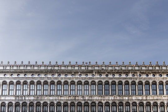 View of the historic Procuratie Nuove building with its repetitive arched windows and ornate facade under a clear blue sky in Venice, Veneto, Italy.