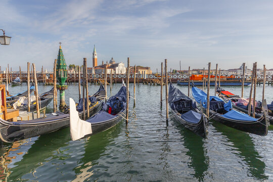 View of traditional gondolas moored to wooden poles in the calm water of the lagoon with historic buildings in the background in Venice, Veneto, Italy.
