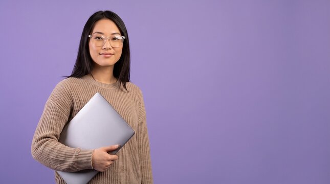 Young Asian woman wearing glasses and a sweater holding a laptop computer against a purple background