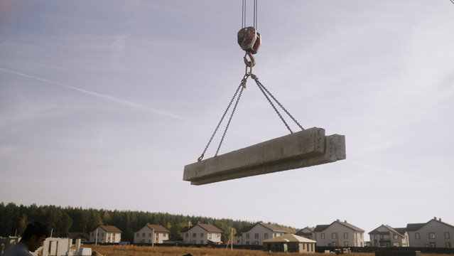 Construction crane lifting heavy concrete beams with chains over new suburban houses, residential building development