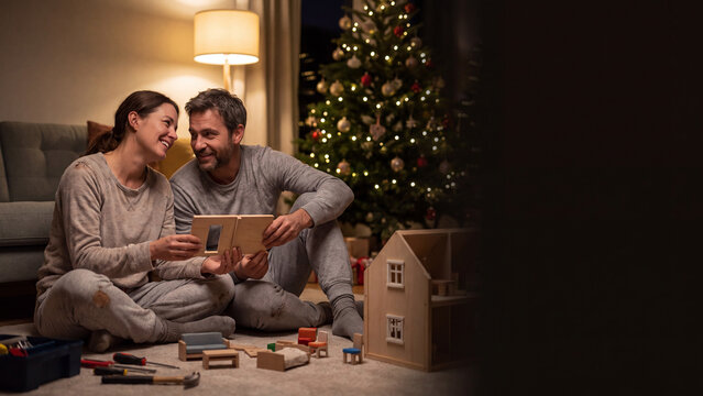 Parents quietly assembling wooden dollhouse late at night with blurred Christmas tree background banner.