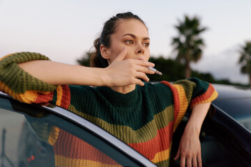 Obraz premium Young woman smoking cigarette leaning on car door outdoors. Casual style and thoughtful mood. Concept of bad habits, lifestyle, and urban youth culture. Soft natural lighting creates a calm