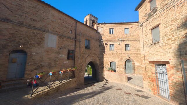 Scorcio di una piazza in un borgo medievale con antichi edifici in pietra, arco pedonale, vicolo e campanile sullo sfondo in una giornata soleggiata