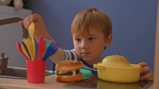 Boy lifts toy burger bun with both hands and stacks pretend burger on toy plate at play kitchen in building; playfulness imagination.