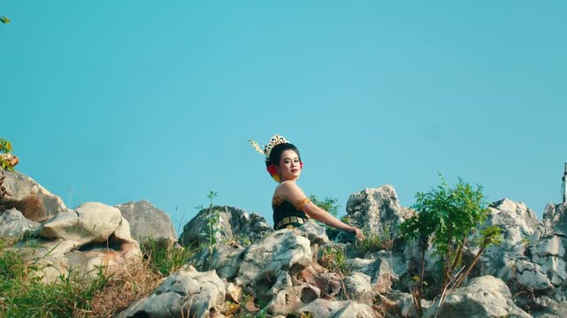Medium portrait of a beautiful Indonesian model wearing a green and gold Kebaya, sitting comfortably on white stone rocks and looking at the camera.