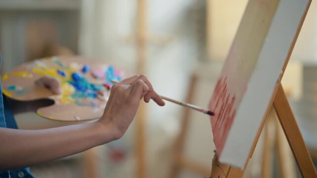 Gen Z Asian woman freelance artist painting with paintbrush on canvas easel, representing creative professional women designer, online independent creator and art studio hobby lifestyle