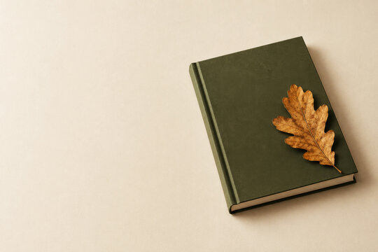 dark green hardcover book with single dried oak leaf resting on neutral beige background