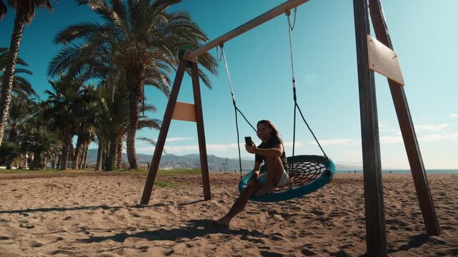 Handsome young man with long hair smiling while holding a smartphone on a swing in a sunny beachside park with palm trees and ocean views, swing set, travel lifestyle, digital nomad